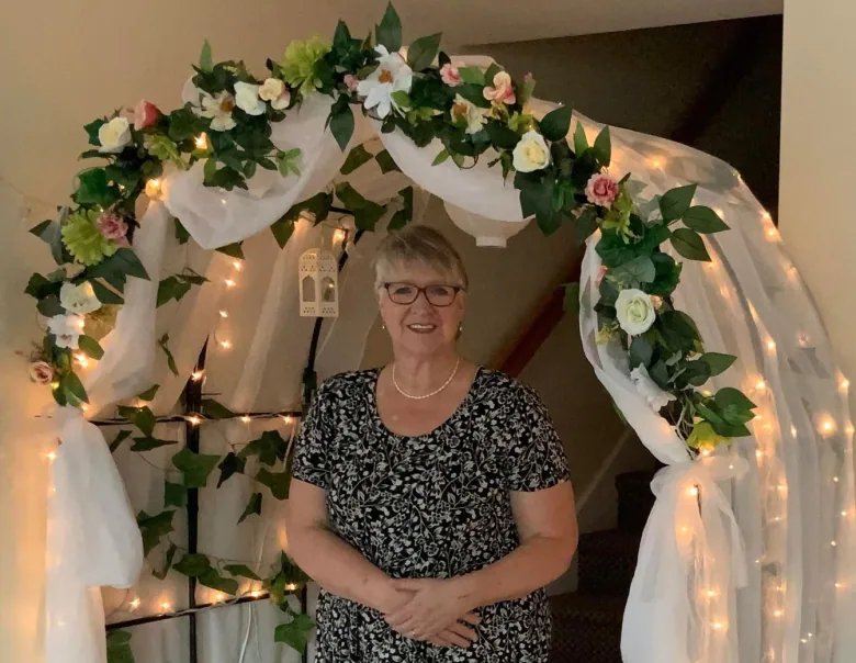 Woman stands under decorated wedding arch with flowers, lights and white tulle.